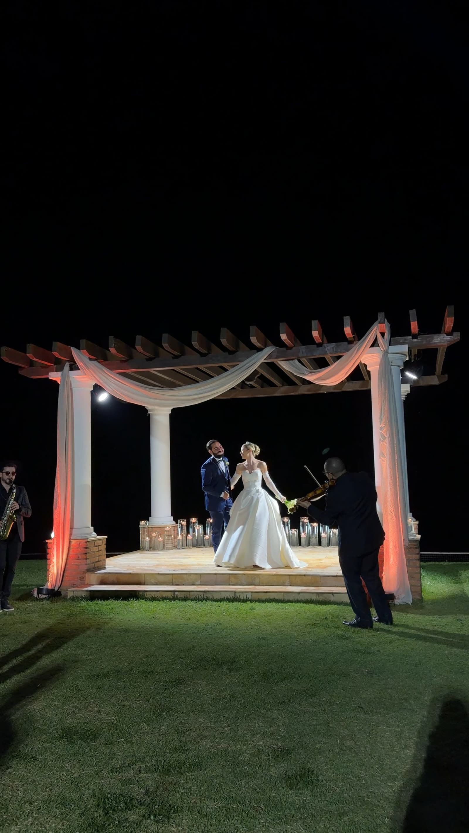 Noivos de mãos dadas sob uma pérgola de tecido iluminada por velas, à noite, com violinista e saxofonista ao lado.